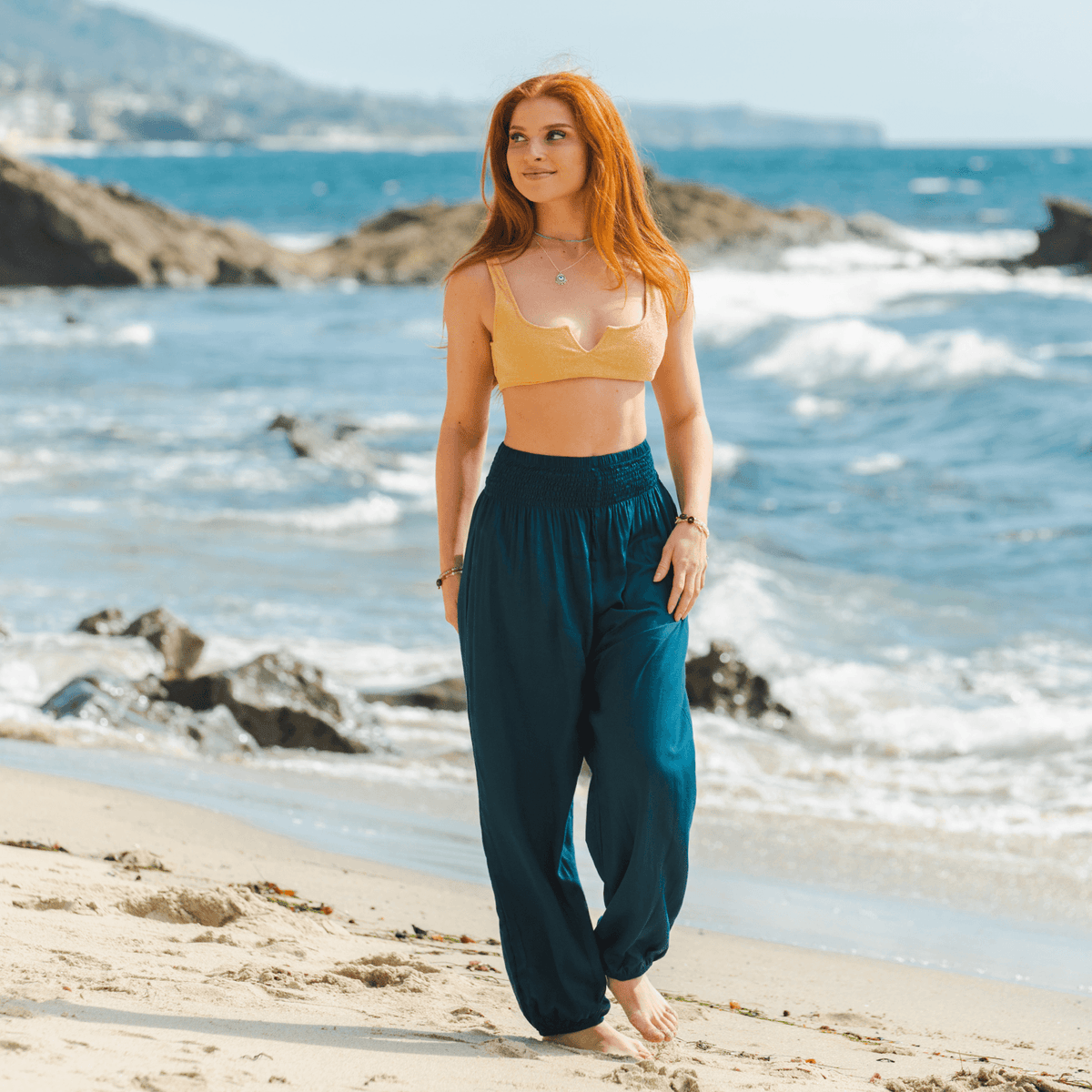 A girl walking by the ocean wearing deep teal harem pants