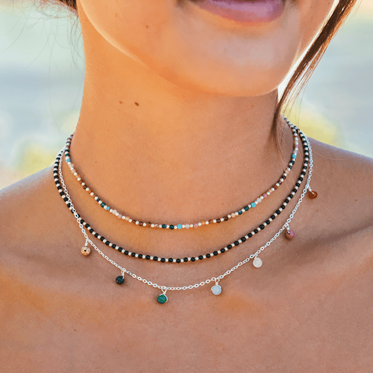 Model wearing a stack of three necklaces. One is a dewdrop charm necklace with multicolor stones and a gold chain, the other is a black stone and gold bead necklace and the last one is a dainty multicolor stone and gold bead necklace